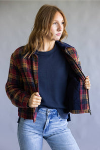 A woman wearing a red and navy plaid flannel shirt jacket over a navy ribbed knit sweater and light wash blue jeans, standing against a neutral gray studio background.