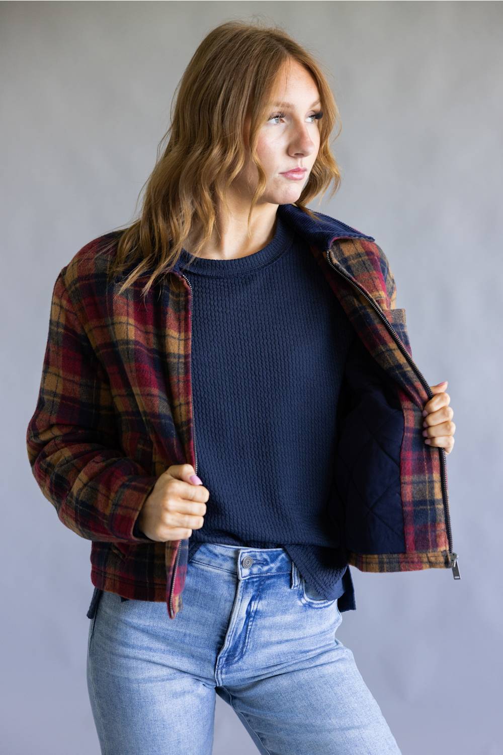 A woman wearing a red and navy plaid flannel shirt jacket over a navy ribbed knit sweater and light wash blue jeans, standing against a neutral gray studio background.