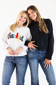 Two women posing together against a white studio background. The woman on the left wears a white sweatshirt with a colorful 'thankful' graphic and blue jeans, while the woman on the right wears a black button-up cardigan and blue jeans.