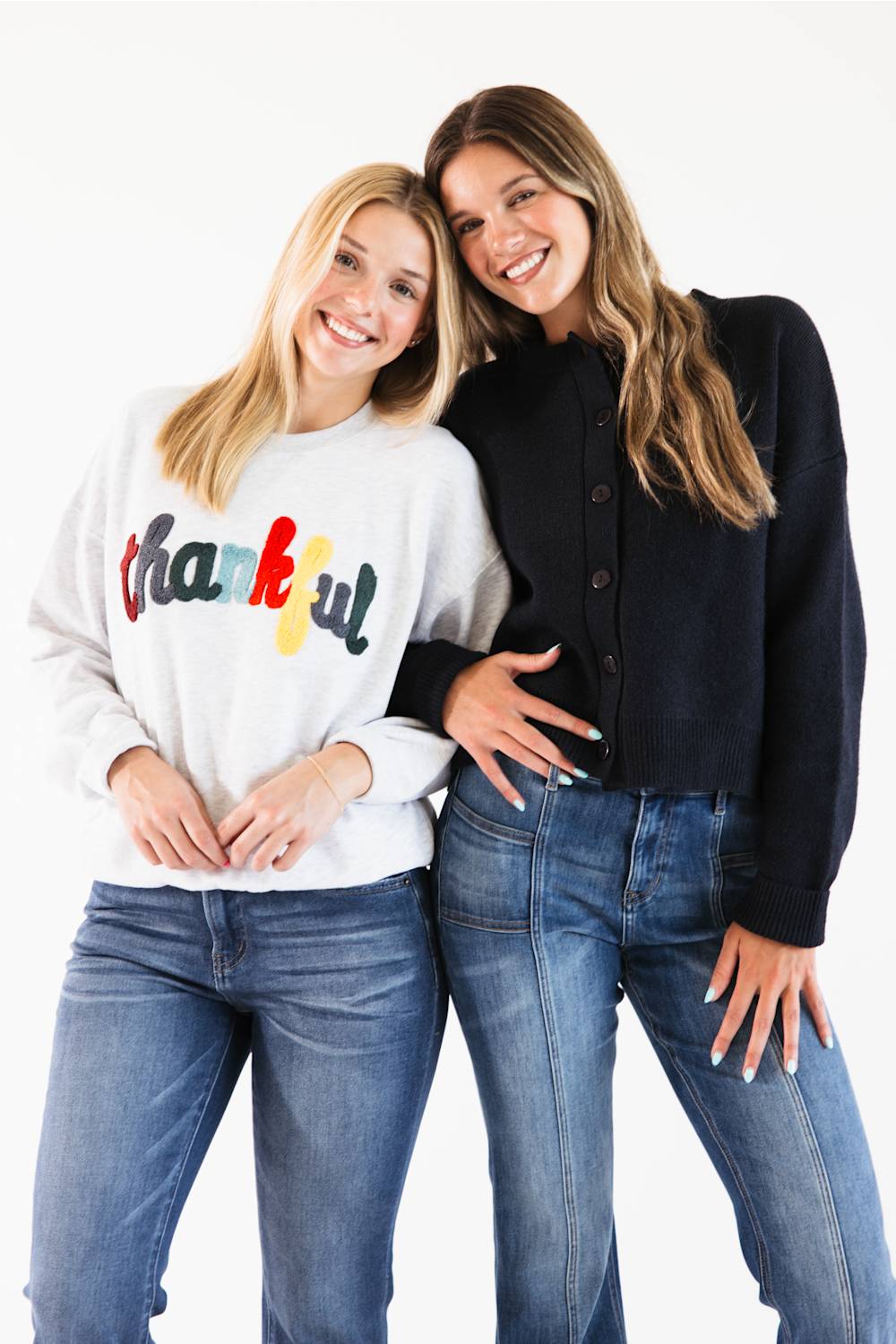 Two women posing together against a white studio background. The woman on the left wears a white sweatshirt with a colorful 'thankful' graphic and blue jeans, while the woman on the right wears a black button-up cardigan and blue jeans.