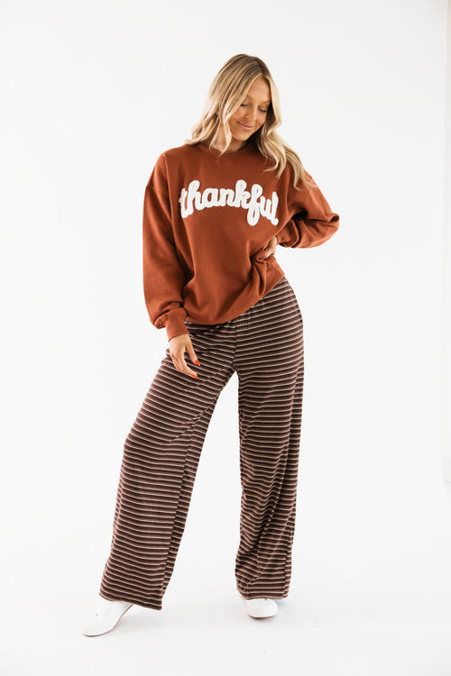 A woman modeling a rust brown sweatshirt with 'thankful' printed in white script across the chest, paired with brown and black horizontally striped wide-leg pants, against a plain white studio background.
