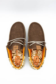 A pair of brown boat-style slip-on shoes with white laces, featuring a patterned orange and beige insole and a small tan brand patch on the side, photographed from above on a white background.