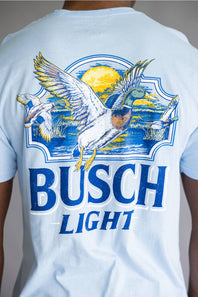 A man wearing a light blue crewneck t-shirt with a large graphic on the back. The graphic features the words 'BUSCH LIGHT' in blue, a white goose with yellow wings, and a yellow sun over blue waves.