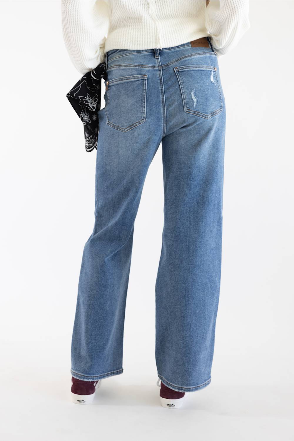 A person shown from the back wearing light blue wide-leg jeans, a white ribbed sweater, and dark shoes, with a black patterned clutch tucked behind the hip, against a plain white studio background.