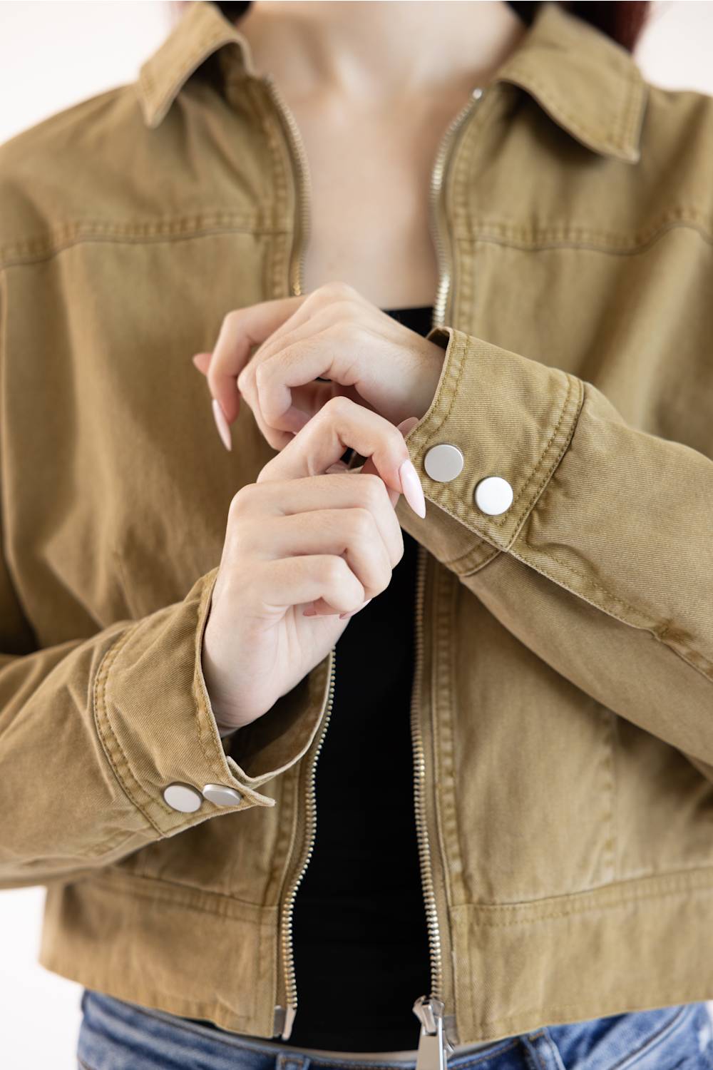 A close-up of a person wearing a tan utility jacket with silver snap buttons on the cuffs and a front zipper, layered over a black top and blue jeans.