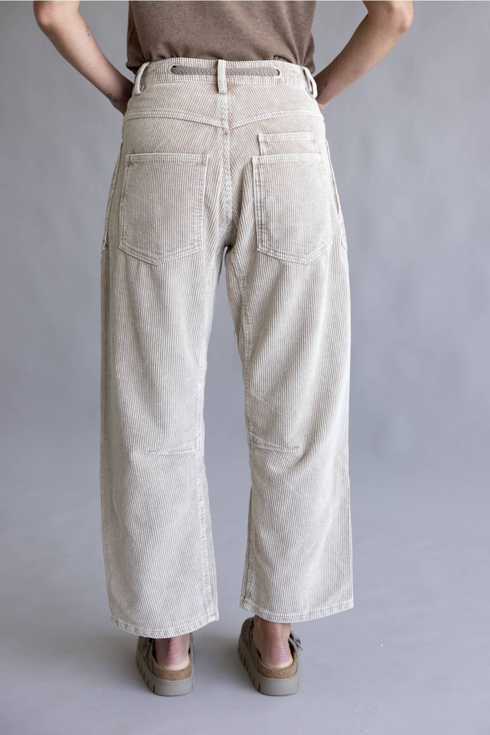 A person shown from the back wearing light beige wide-leg corduroy pants with a belt and back pockets, paired with brown sandals against a plain gray studio background.