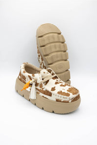 A pair of platform mule shoes featuring a brown and cream cow-print pattern on the upper, a beige platform sole, and a white tassel charm with an orange accent, shown against a plain white background.