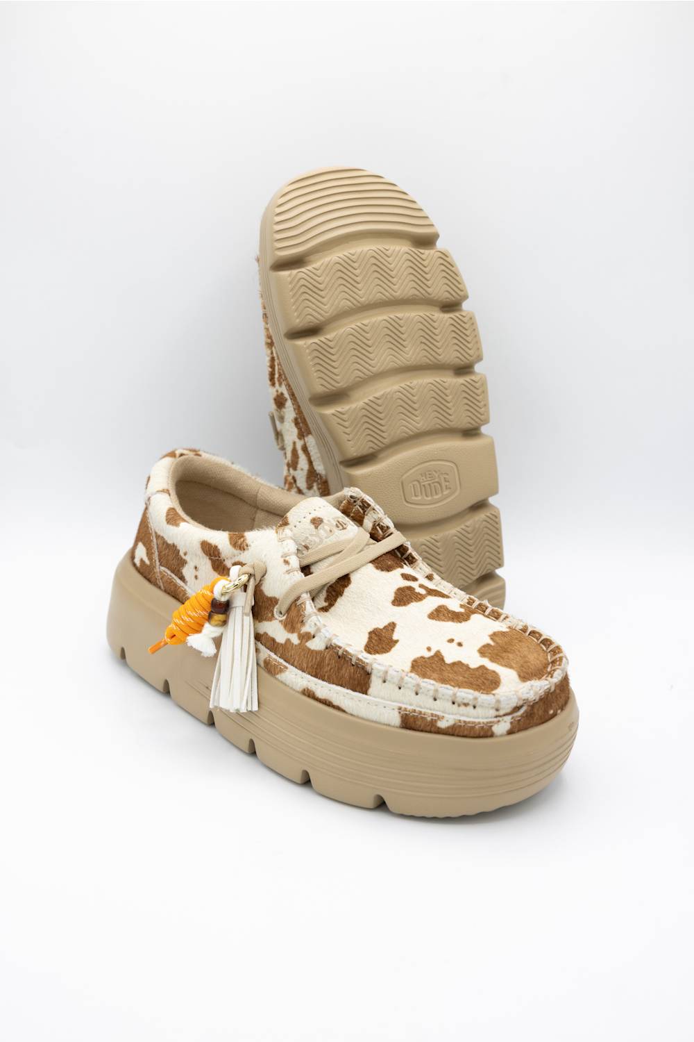 A pair of platform mule shoes featuring a brown and cream cow-print pattern on the upper, a beige platform sole, and a white tassel charm with an orange accent, shown against a plain white background.