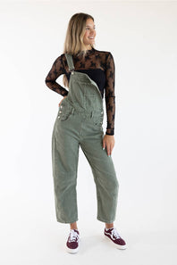 A woman modeling olive green corduroy bib overalls over a black sheer long-sleeve top and black pants, paired with burgundy sneakers, against a plain white studio background.