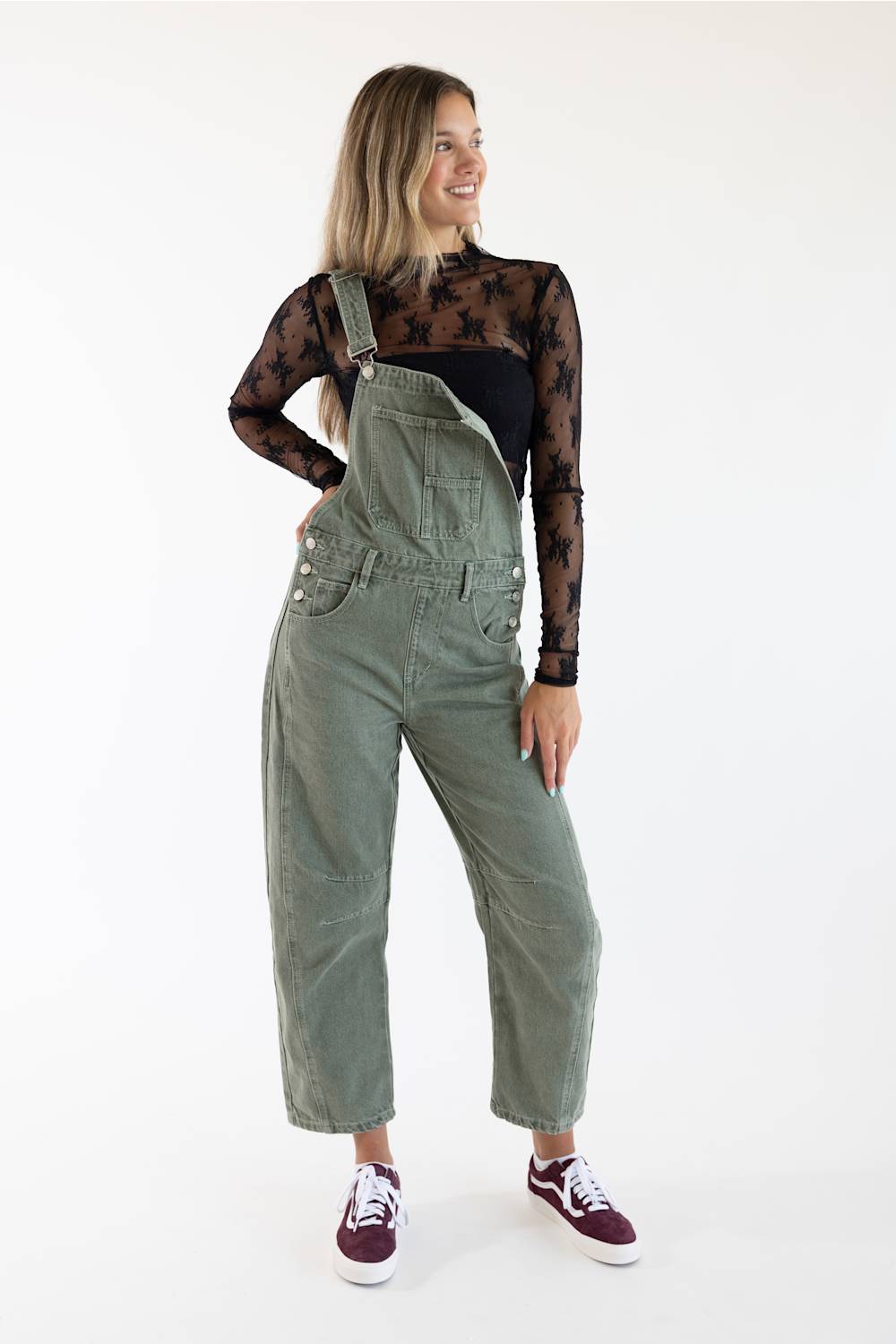 A woman modeling olive green corduroy bib overalls over a black sheer long-sleeve top and black pants, paired with burgundy sneakers, against a plain white studio background.