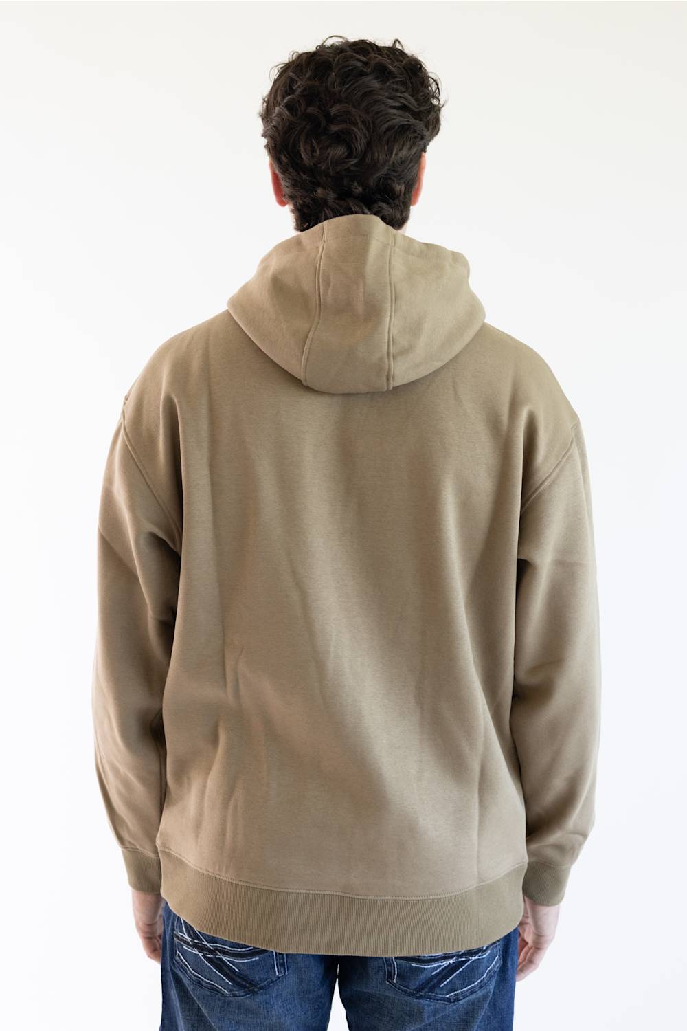 A man wearing a tan pullover hoodie with 'ARIAT' embroidered vertically in brown on the right sleeve and a small yellow logo patch near the cuff, paired with blue jeans against a plain white background.