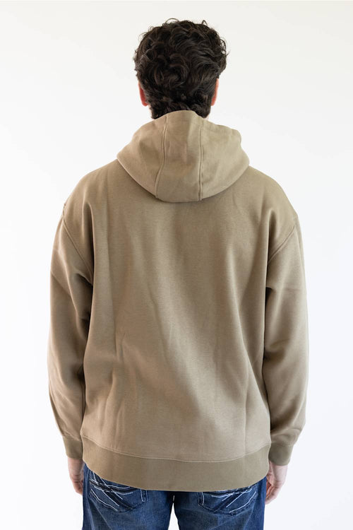 A man wearing a tan pullover hoodie with 'ARIAT' embroidered vertically in brown on the right sleeve and a small yellow logo patch near the cuff, paired with blue jeans against a plain white background.
