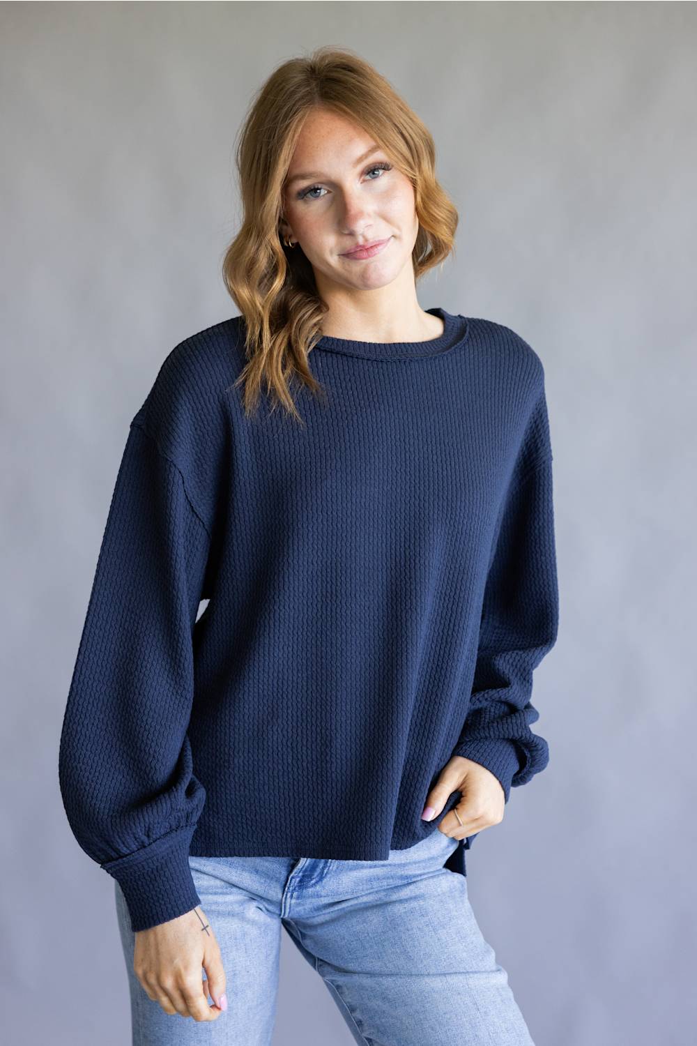 A woman wearing a navy blue waffle-knit crewneck sweater and light blue jeans, standing against a neutral gray studio background.