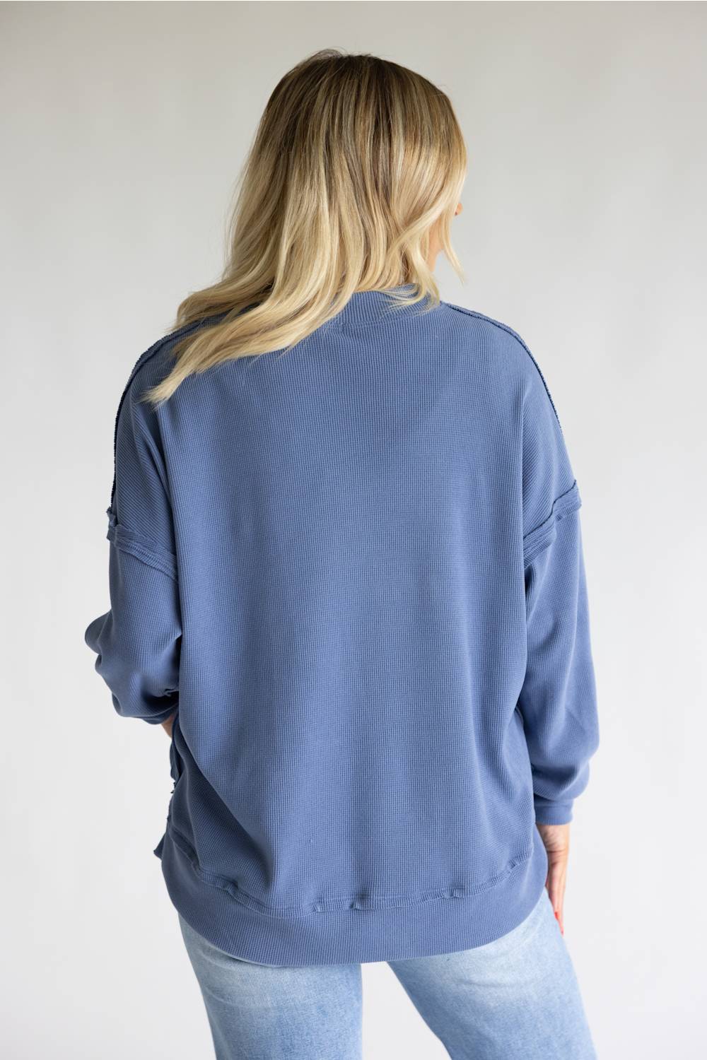 A woman viewed from behind wearing a dusty blue crewneck sweatshirt and light wash jeans against a plain light gray background.
