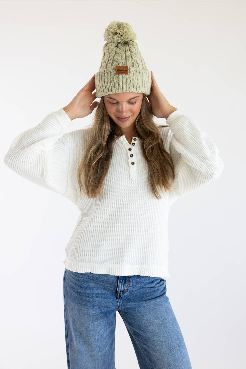 A woman wearing a cream ribbed knit henley sweater, blue jeans, and a light sage green knit beanie with a pom-pom and a small brown leather patch. She is adjusting the beanie against a plain white studio background.