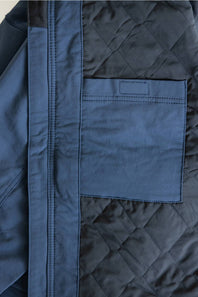 Close-up of a navy blue quilted jacket showing a diagonal seam, a solid fabric panel, and a stitched chest pocket.