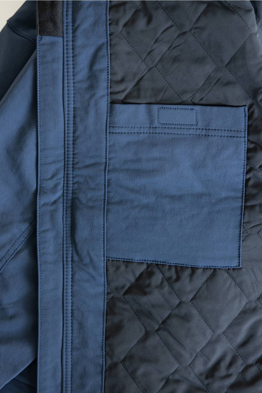 Close-up of a navy blue quilted jacket showing a diagonal seam, a solid fabric panel, and a stitched chest pocket.