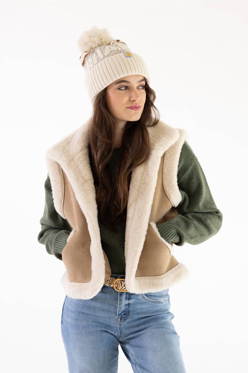 Close-up front view of a model looking down while wearing a Carhartt thick knit cuffed beanie with a pom-pom in Oat Milk and a beige sherpa vest