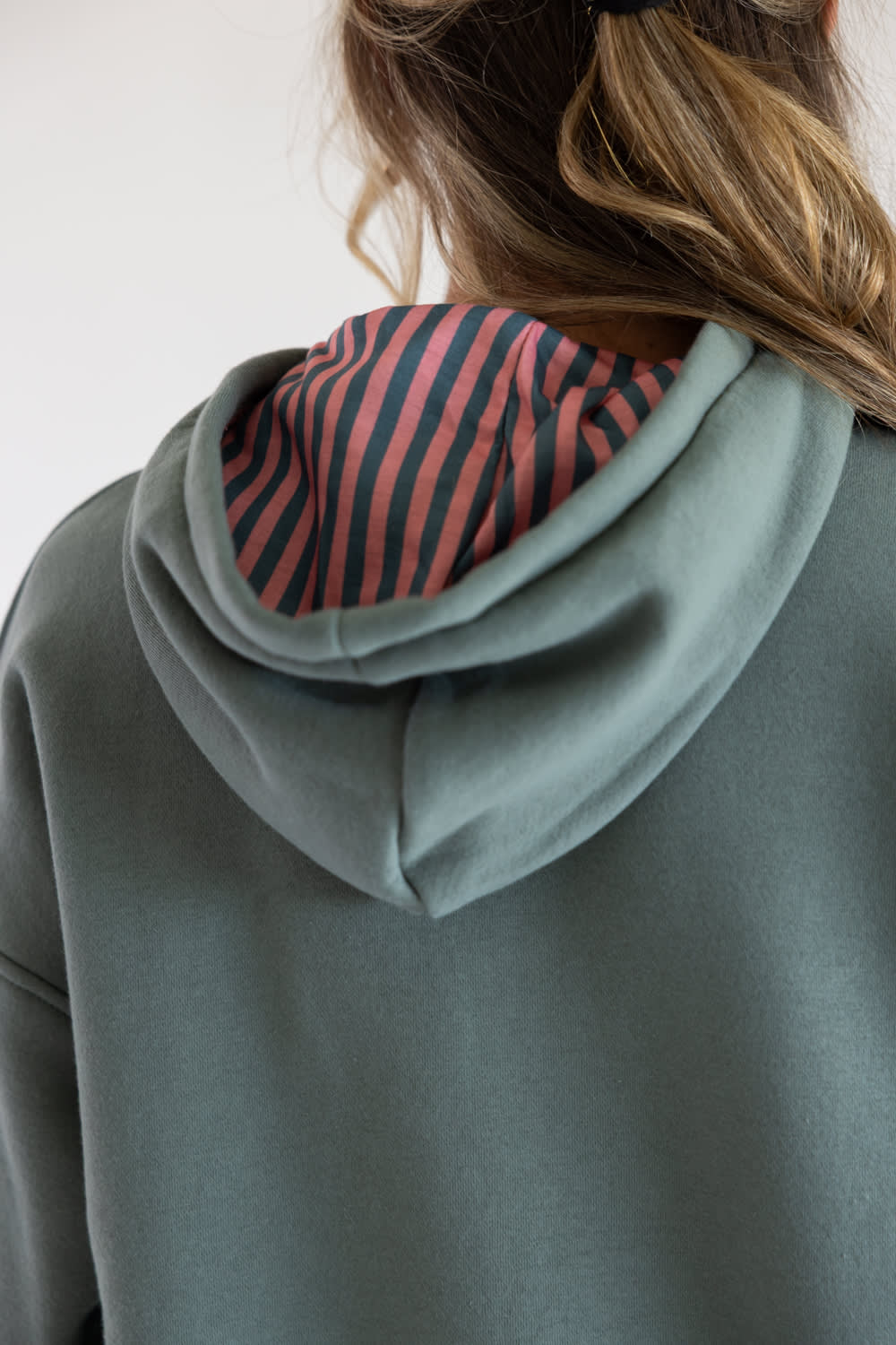 Close-up back view of a person wearing a sage green grey   hoodie with a pink and navy striped hood lining.