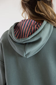 Close-up back view of a person wearing a sage green grey   hoodie with a pink and navy striped hood lining.
