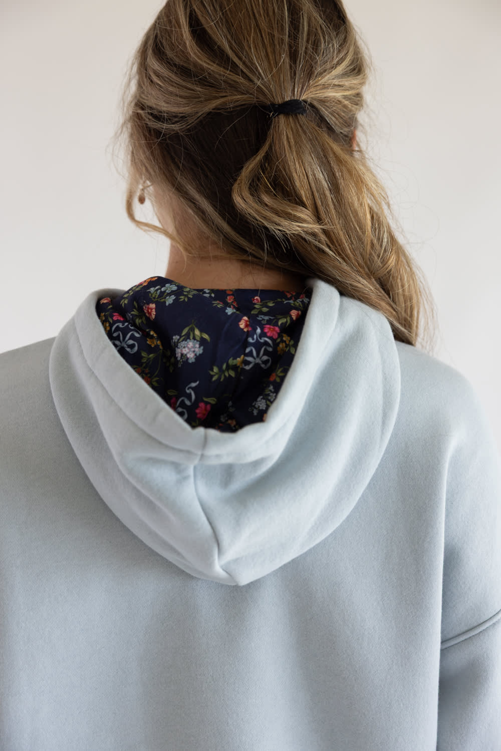 Back view of a person wearing a light gray hoodie with a dark navy floral lining inside the hood, set against a plain off-white background.