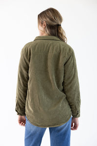 A woman viewed from behind wearing an olive green corduroy button-up shirt and blue jeans against a plain white background.