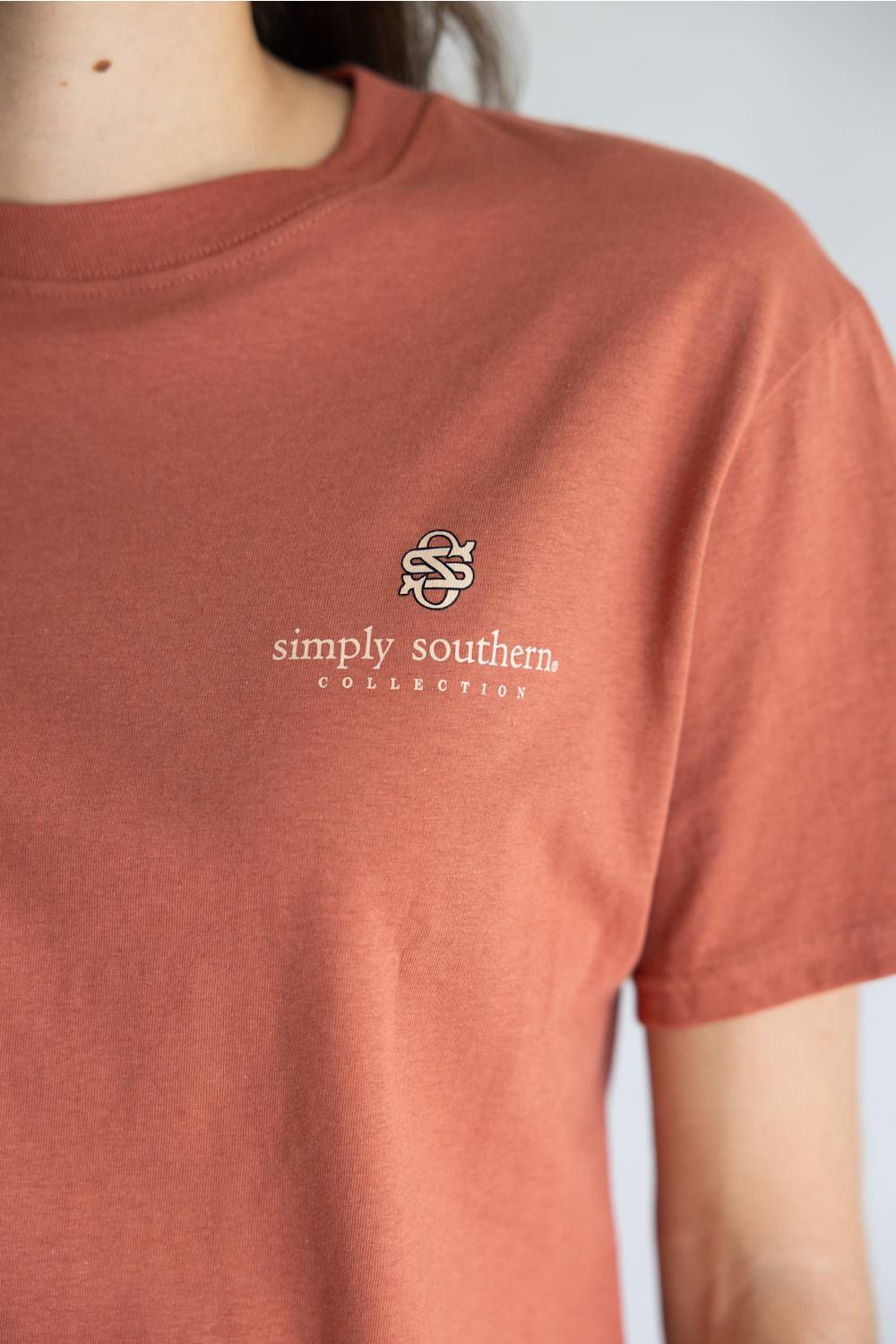 A close-up of a person wearing a terracotta-colored short-sleeve t-shirt with a small white  logo and the text 'simply southern COLLECTION' on the left chest.