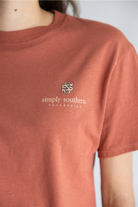 A close-up of a person wearing a terracotta-colored short-sleeve t-shirt with a small white  logo and the text 'simply southern COLLECTION' on the left chest.