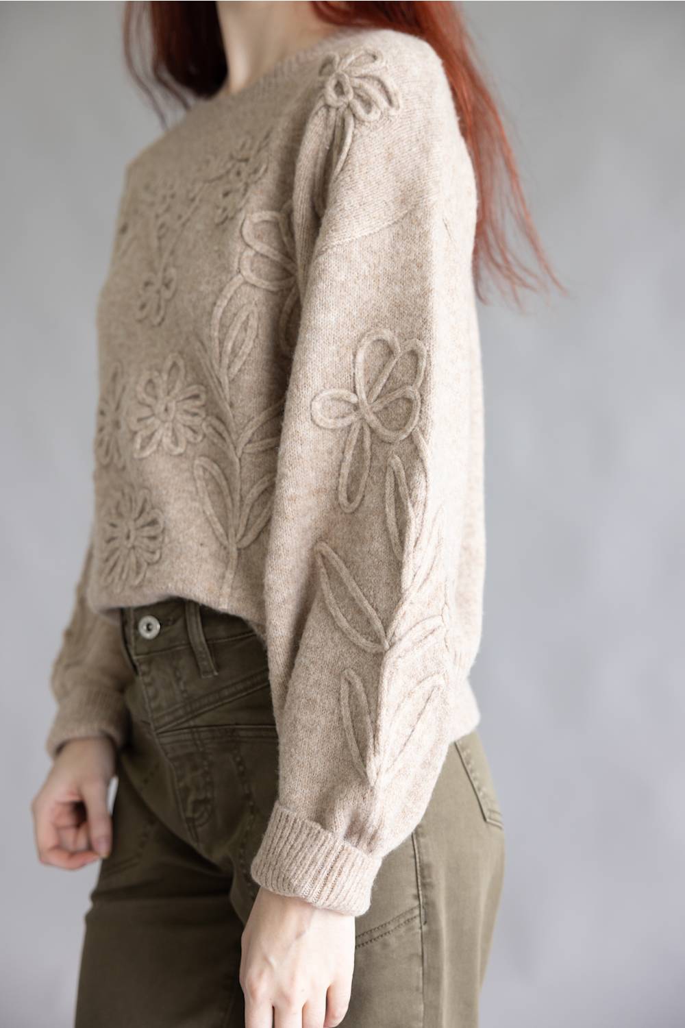 A person wearing a cropped beige knit sweater with raised floral and leaf patterns on the sleeves and chest, paired with olive green pants, against a plain light gray background.