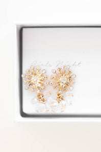 A pair of gold snowflake-shaped stud earrings with clear rhinestones and clear tassel drops, presented inside a white gift box with 'Rains Night' printed on the insert.