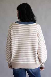 A person shown from the back wearing a cream and beige horizontally striped knit sweater with a contrasting blue ribbed crew neck, paired with blue jeans, against a neutral gray background.