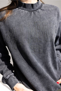 A close-up of a person wearing a charcoal gray long-sleeve mock neck sweatshirt with tonal embossed lettering across the chest.