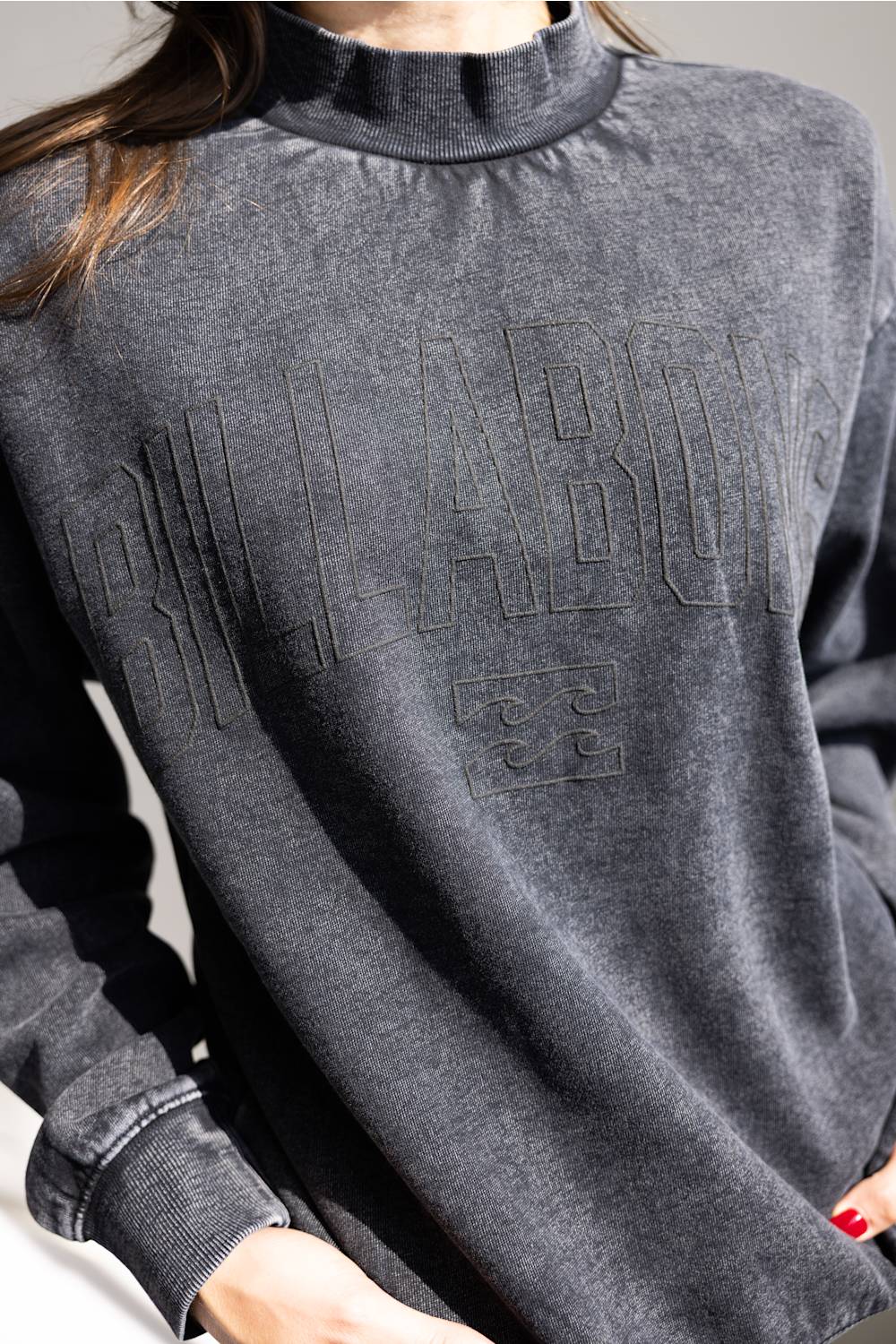 A close-up of a person wearing a charcoal gray long-sleeve mock neck sweatshirt with tonal embossed lettering across the chest.