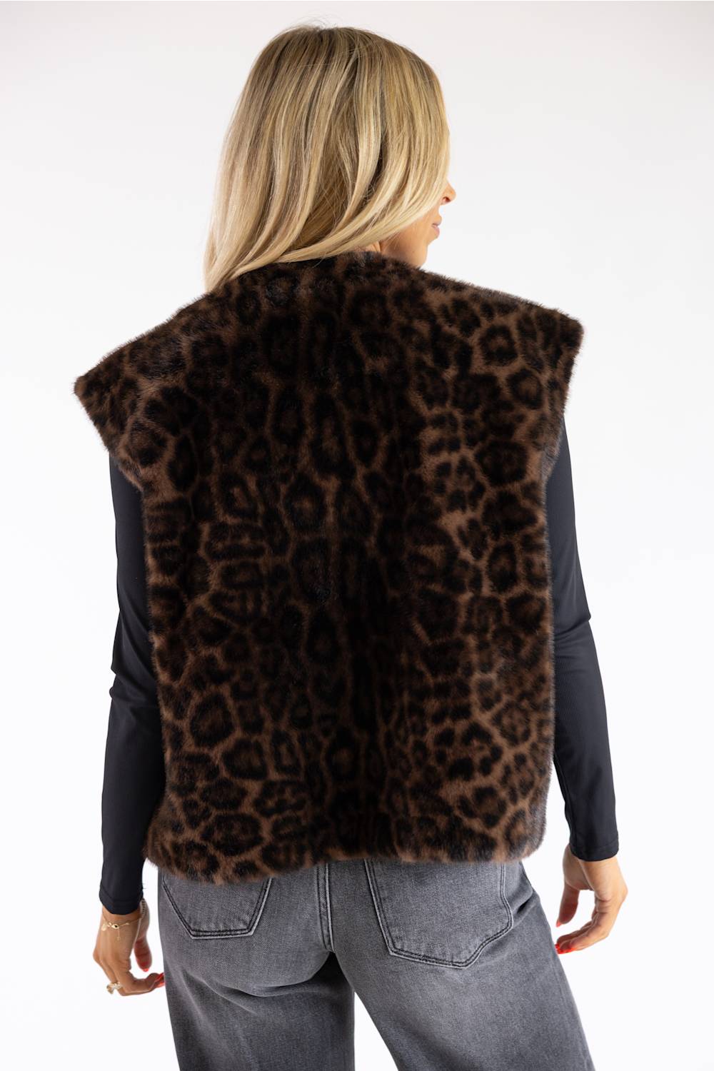 A woman shown from the back wearing a brown leopard-print faux fur vest over a black long-sleeve top and gray jeans, against a plain white background.