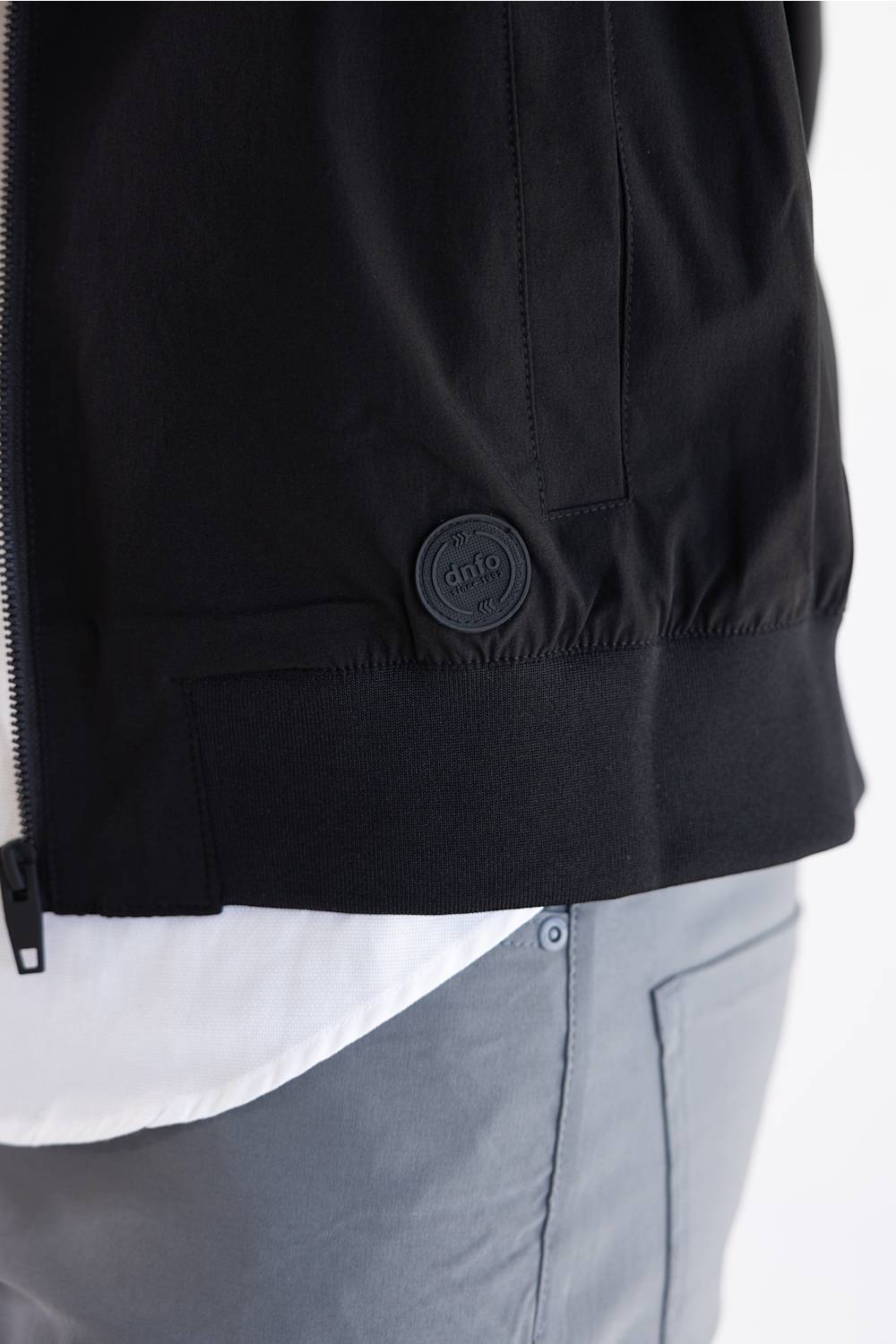 Close-up of a black jacket worn by a model, showing a zippered front, a ribbed hem, and a circular black logo patch on the lower front pocket. A white shirt and gray pants are visible underneath.