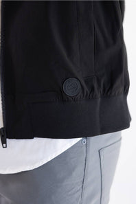 Close-up of a black jacket worn by a model, showing a zippered front, a ribbed hem, and a circular black logo patch on the lower front pocket. A white shirt and gray pants are visible underneath.