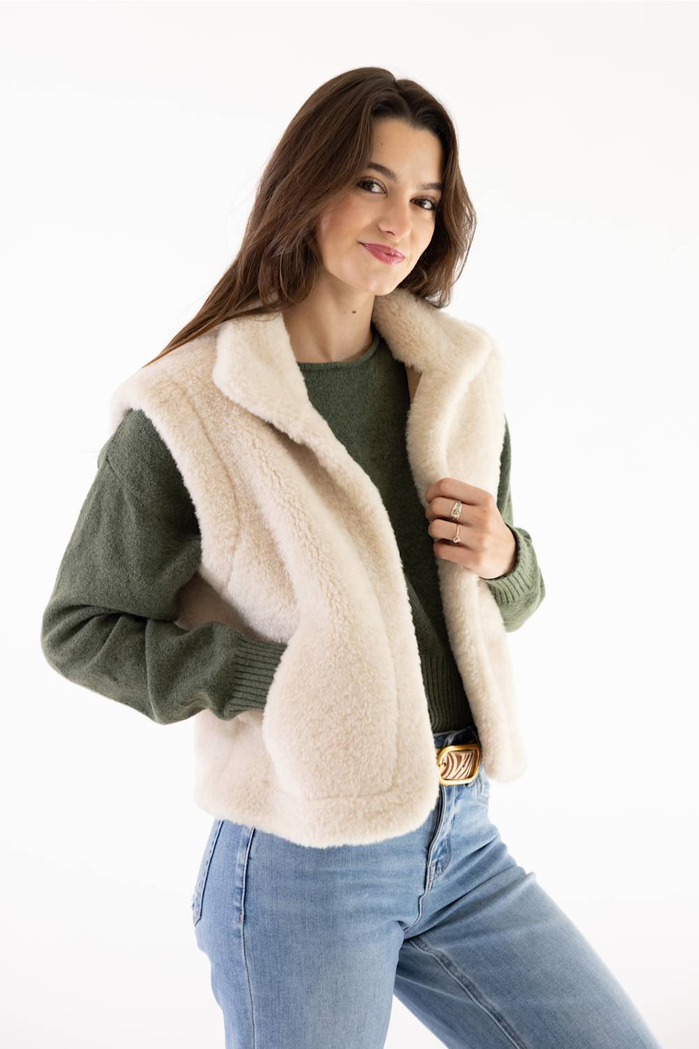 A woman modeling a cream-colored faux fur vest over an olive green sweater and blue jeans, photographed against a clean white background.