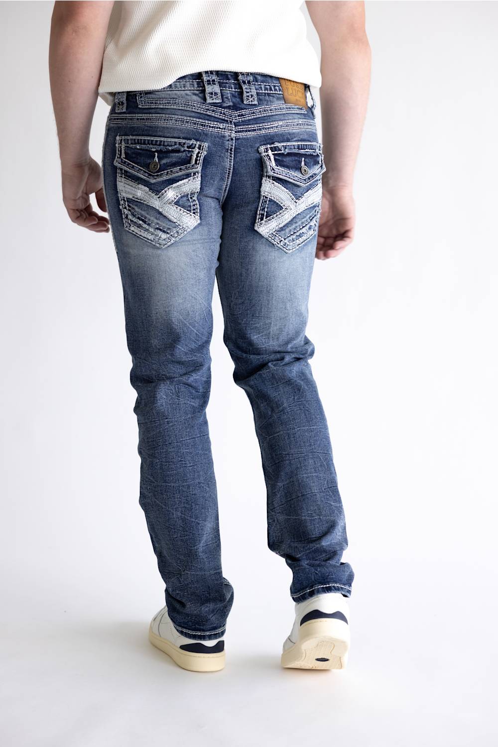 Rear view of a person wearing blue denim jeans with distinctive white contrast stitching and decorative stitching on the back pockets, paired with a white t-shirt and white sneakers.