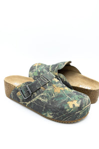 A pair of slip-on clogs with a green and brown woodland camouflage pattern, featuring a cork wedge heel and a black buckle strap across the top, shown on a white background.