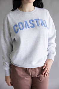 A person wearing a light heather gray crewneck sweatshirt with 'COASTAL' embroidered in blue across the chest, paired with brown pants, against a neutral gray background.