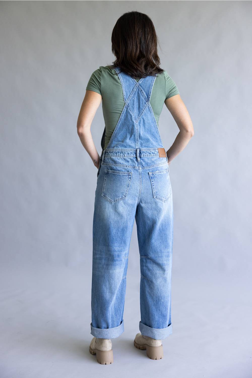A woman viewed from behind wearing light blue denim overall-style pants with a green short-sleeve top and beige shoes, standing against a plain light gray studio background.