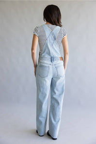 A woman viewed from behind wearing light-wash denim overall over a black and white striped t-shirt, standing against a neutral gray studio background.