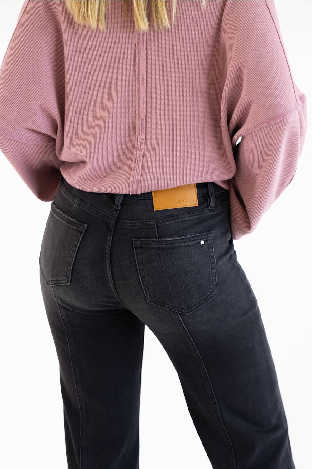 Rear view of a person wearing a dusty pink ribbed long-sleeve top and dark charcoal gray jeans with a tan leather patch at the back waistband, against a plain white background.
