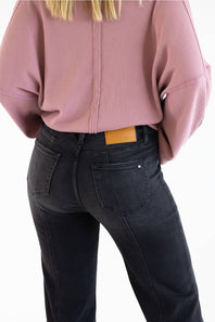 Rear view of a person wearing a dusty pink ribbed long-sleeve top and dark charcoal gray jeans with a tan leather patch at the back waistband, against a plain white background.