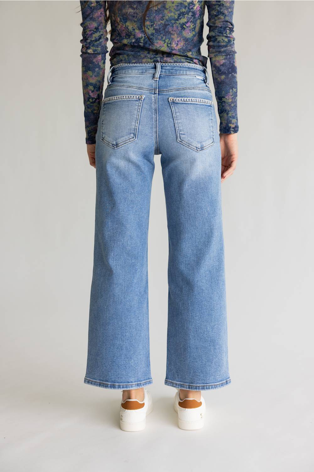 A person shown from the back wearing light-wash blue wide-leg jeans, a dark floral long-sleeve top, and white platform sandals against a plain light gray studio background.