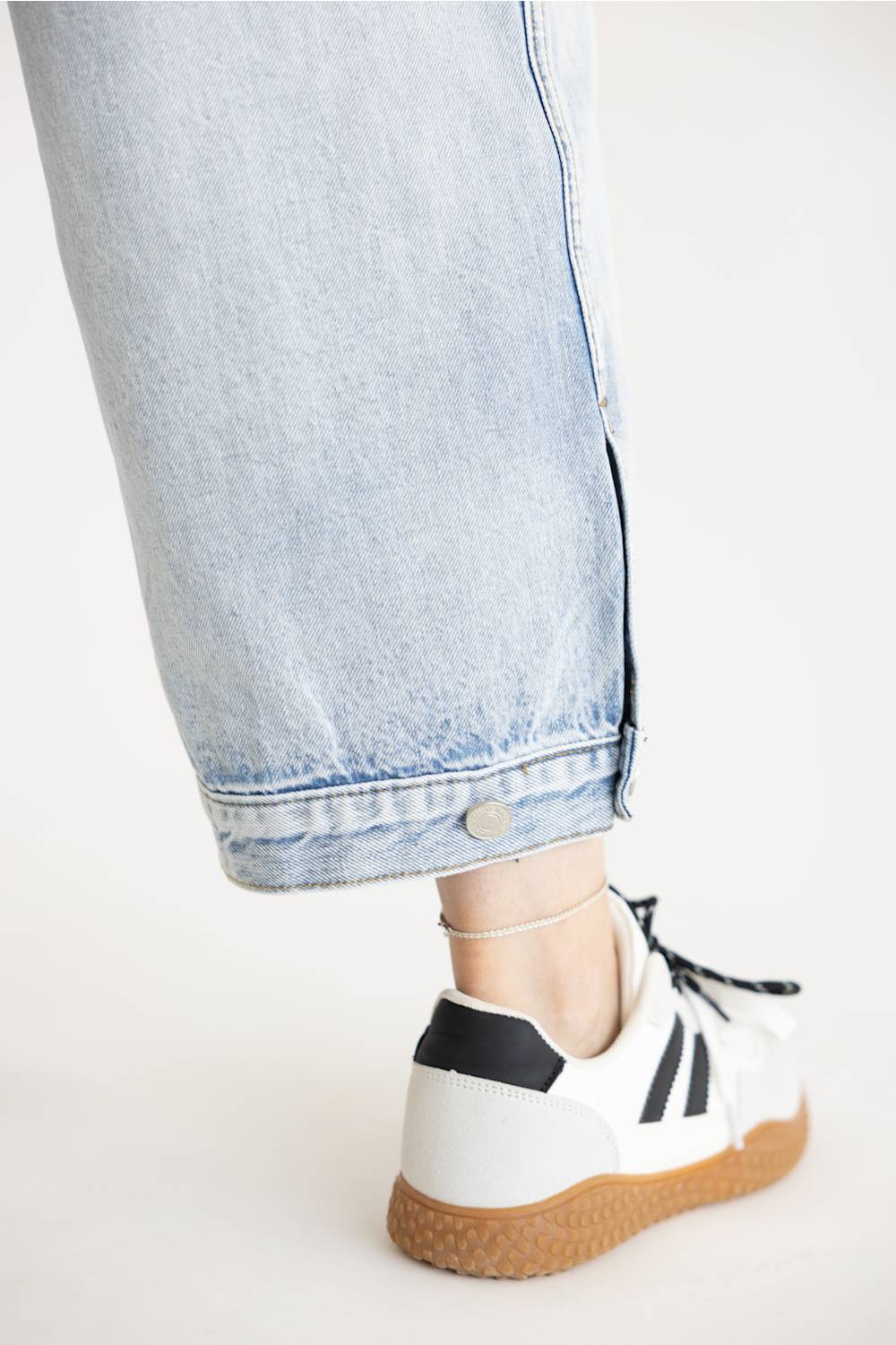 Close-up shot of Aaron & Amber High Rise Sideseam Detail Wide Leg Jeans in Light Denim showing the ankle hem, side button detail, and white-and-brown low-top sneakers.