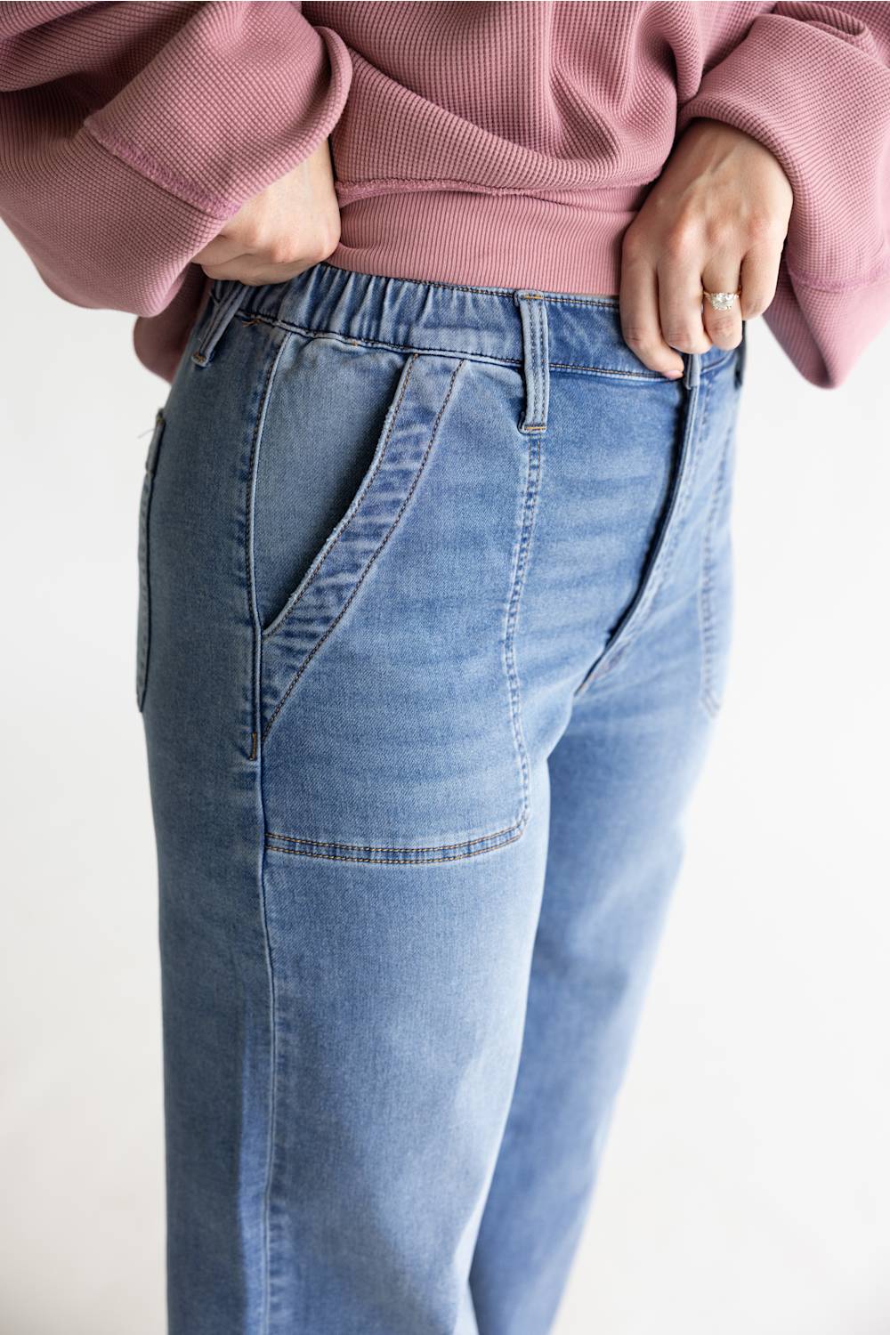 A close-up of a person wearing light blue denim jeans with a pink ribbed sweatshirt, standing against a plain light background.