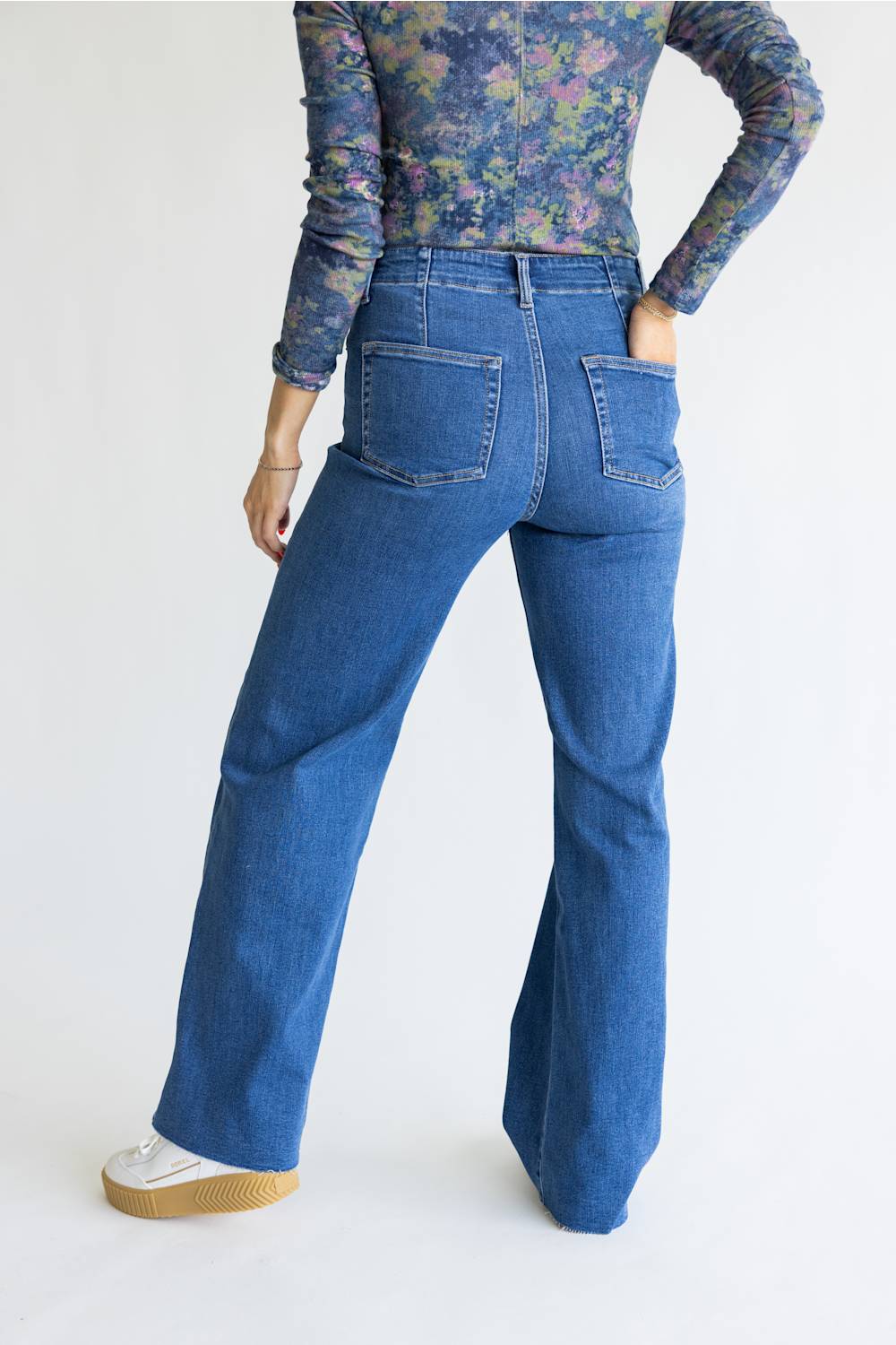 A person shown from the back wearing blue wide-leg jeans, a floral long-sleeve top, and white sneakers against a plain light background.