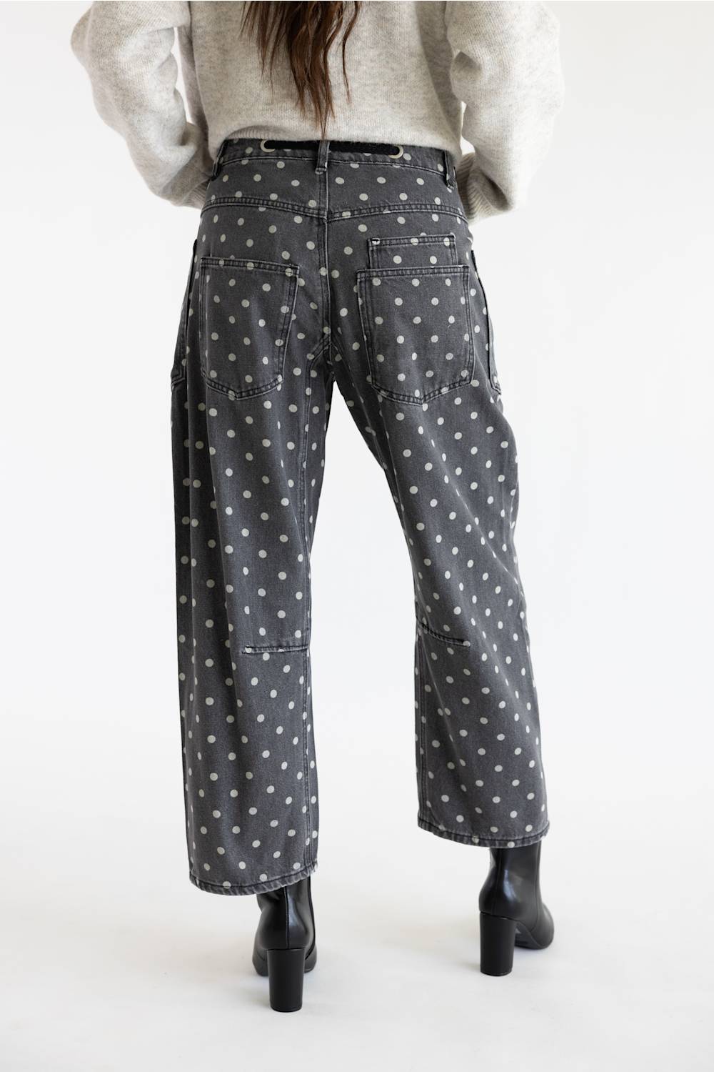 Back view of a model posed from behind wearing black polka dot barrel-leg jeans featuring two rear patch pockets, styled with a beige sweater and black leather heeled boots against a white background.