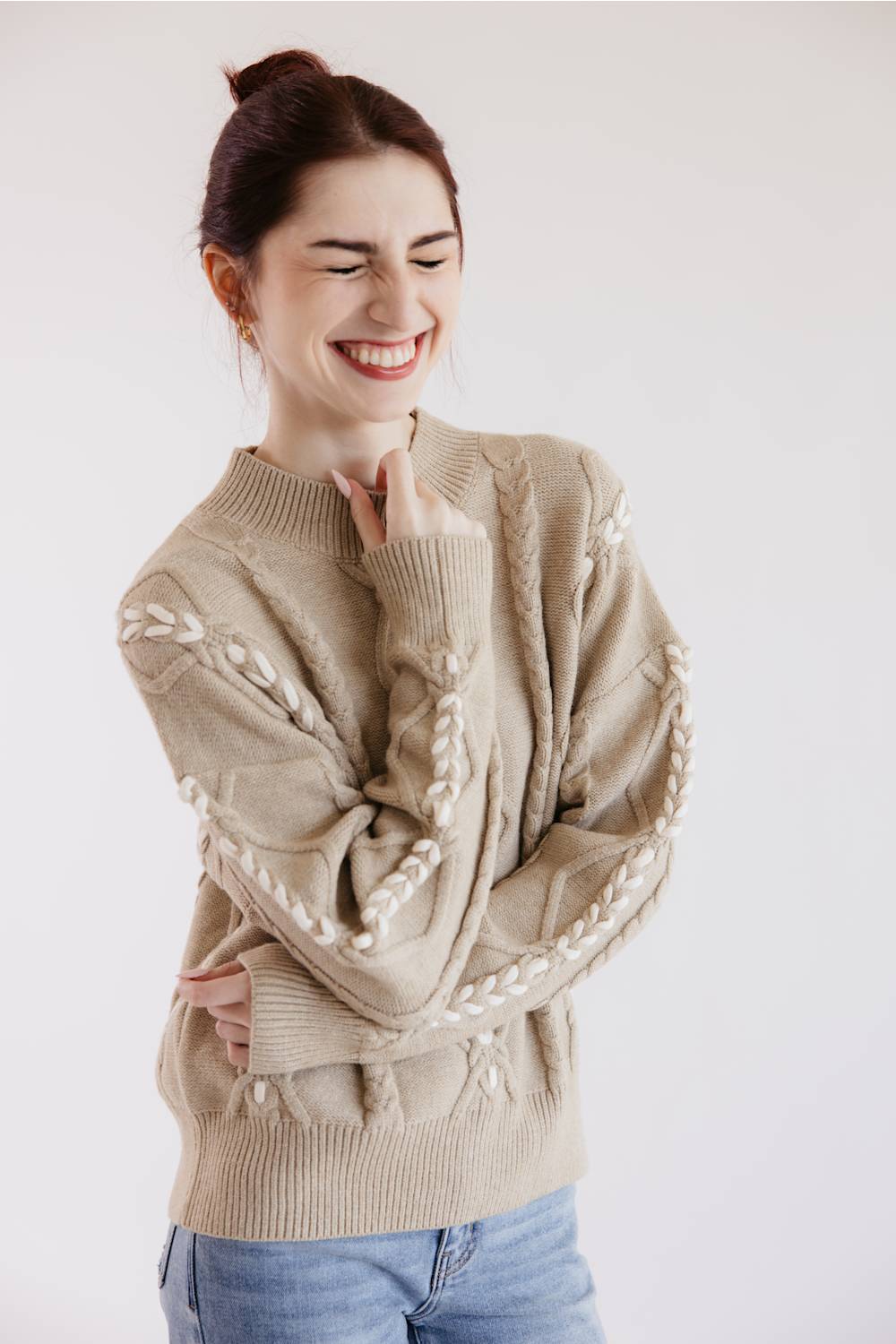 A woman smiling and laughing while wearing a beige cable-knit sweater with white decorative trim, paired with blue jeans, against a plain light background.